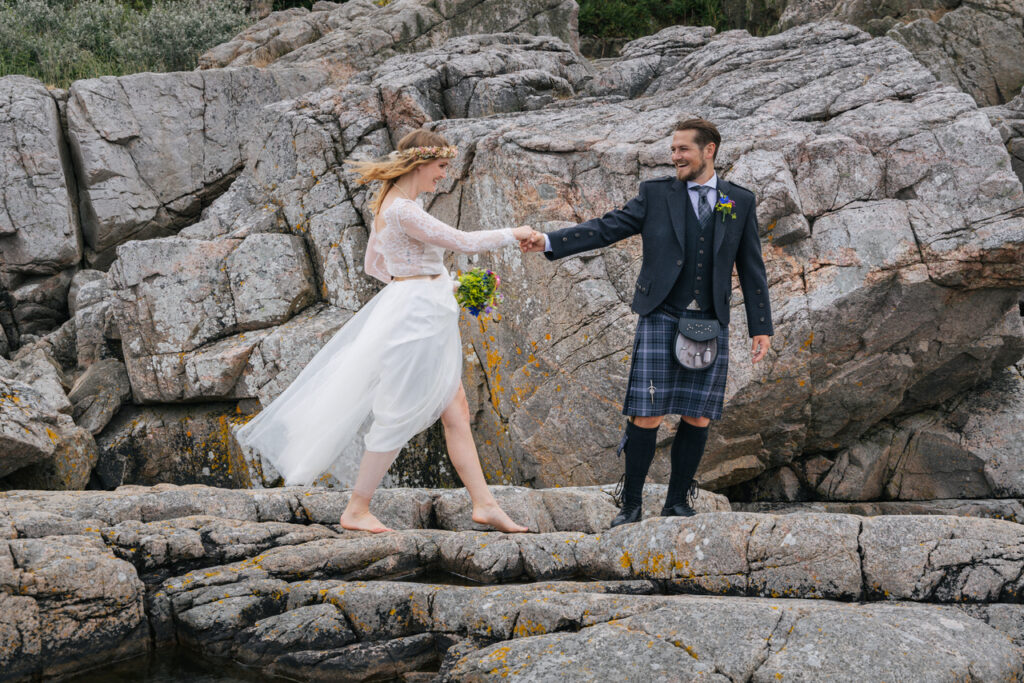 A groom gives hand to the bride, walking on the rocks of Bornholom island, where they have intimate wedding abroad for two.